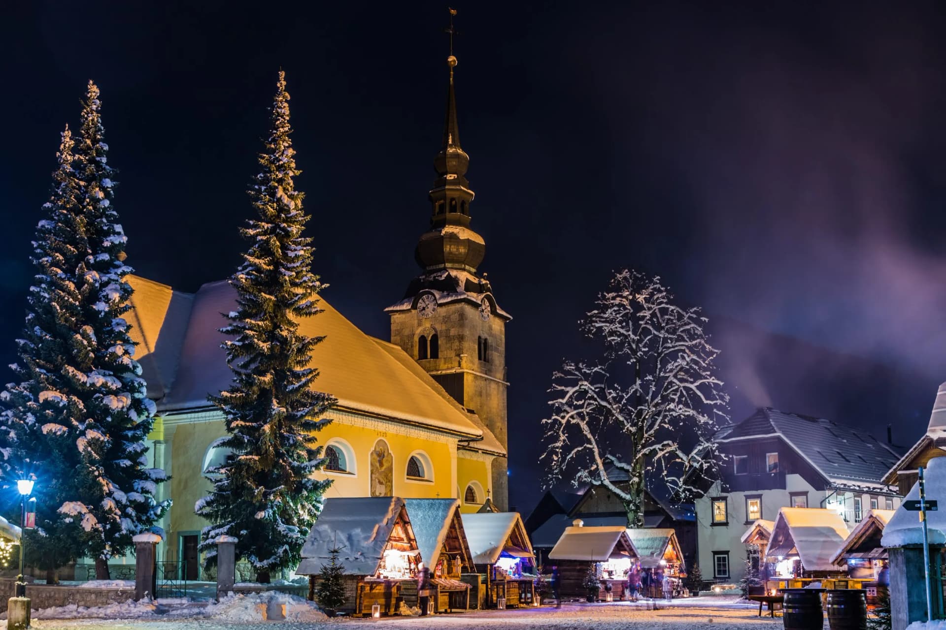 Kranjska Gora winter market with snow-covered church, illuminated stalls, and tall pine trees at night.