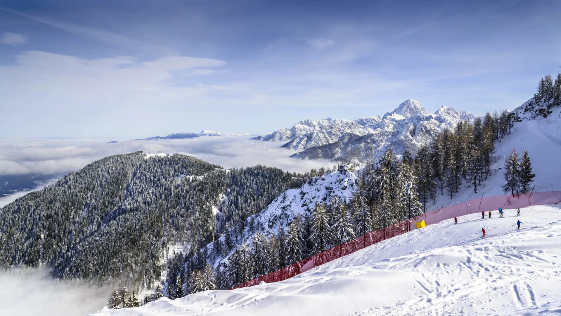 Skiers on snowy slope above clouds with Monte Lussari peaks in background