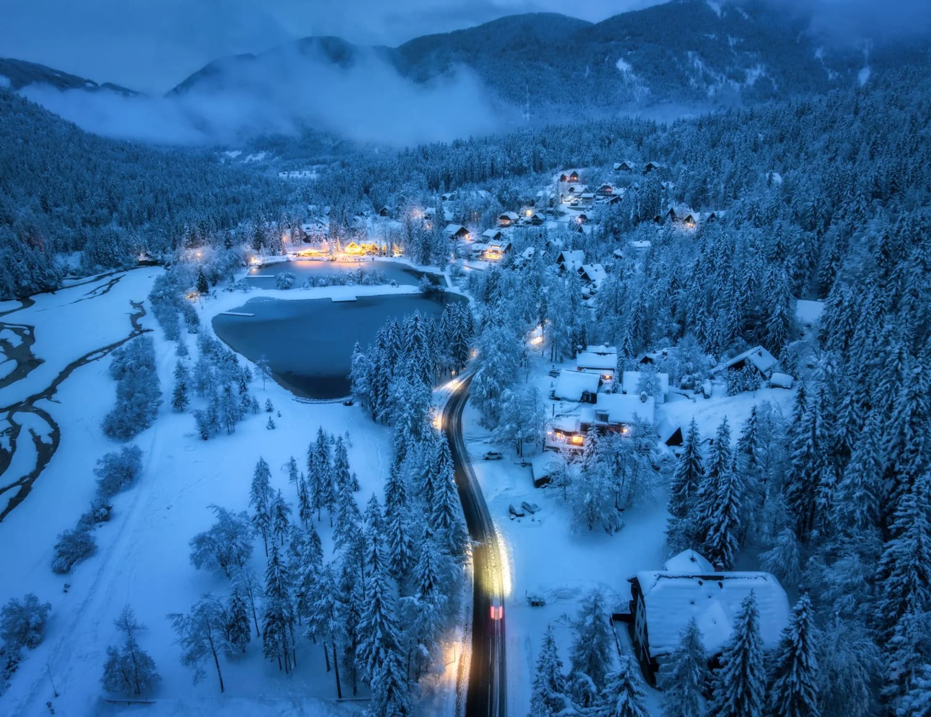 Aerial view of illuminated alpine village with snow-covered trees, frozen lake, and road at dusk.