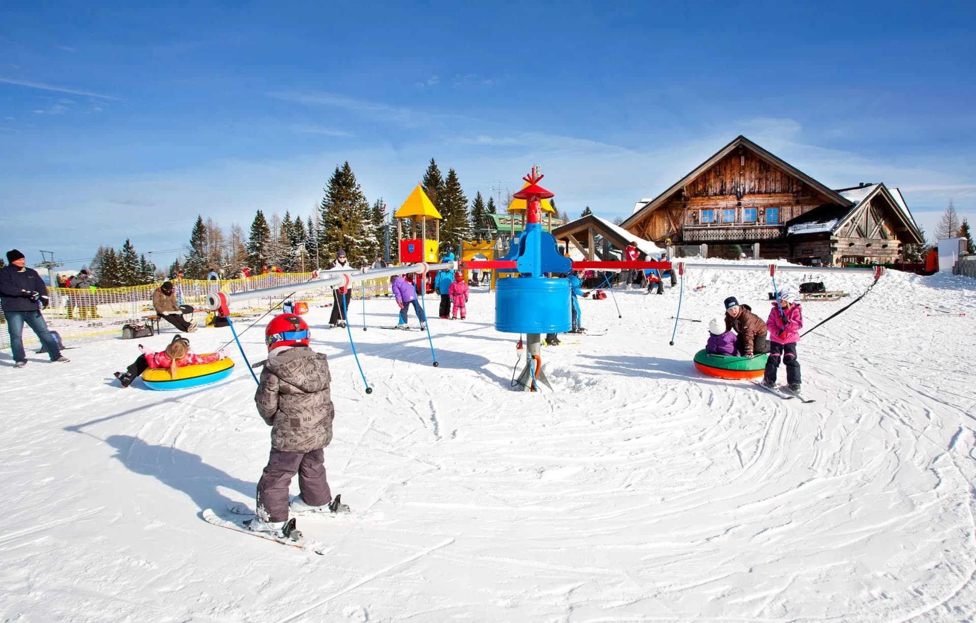 Children skiing and tubing at Cerkno kids park with a wooden lodge in the background.