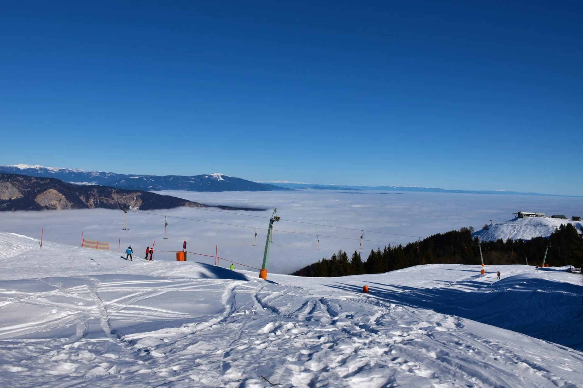 Skiers on snowy slope above cloud inversion with distant snow-capped mountains and clear blue sky.