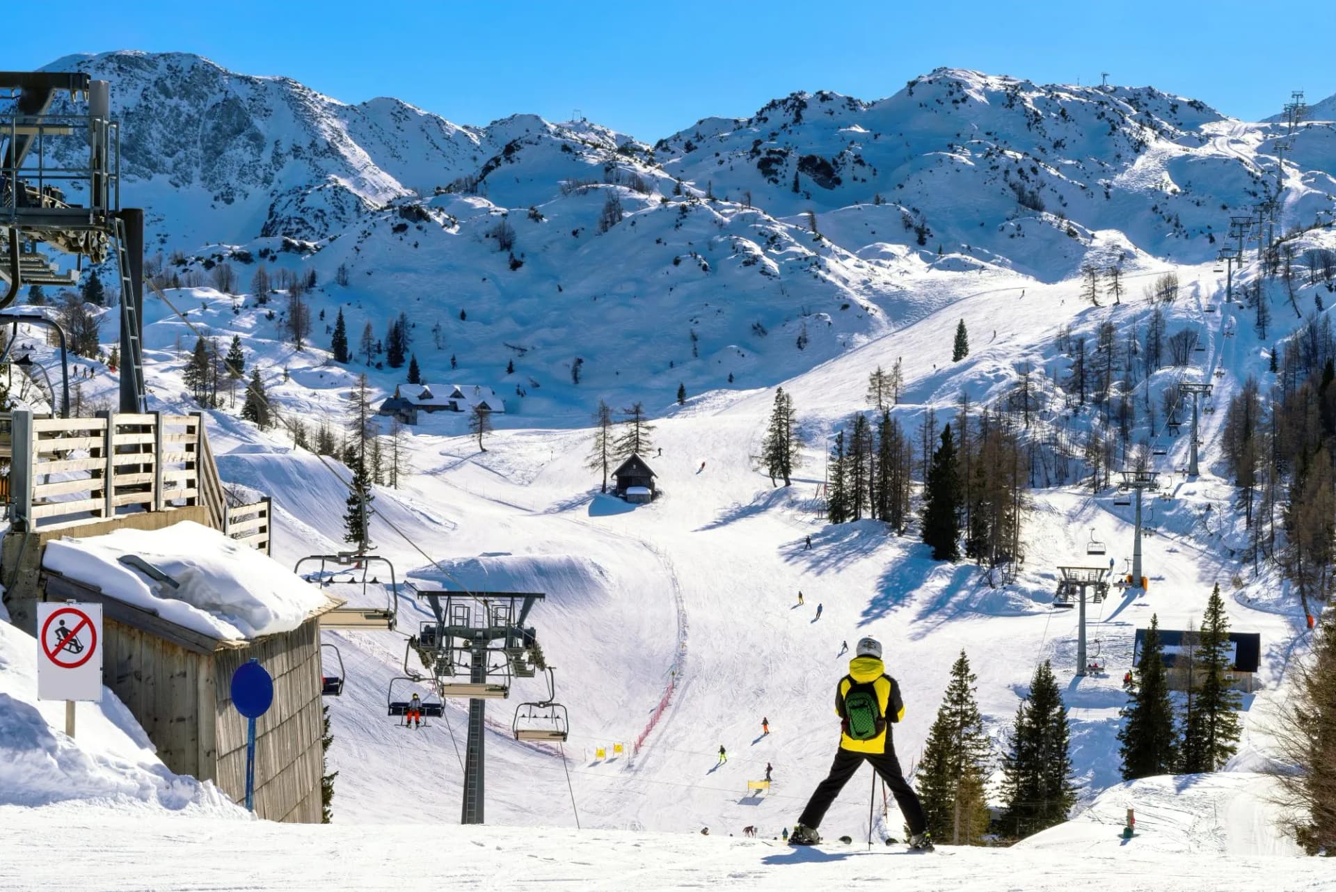 Skiers on snowy slopes with chairlifts and snow-covered mountains under a clear blue sky.