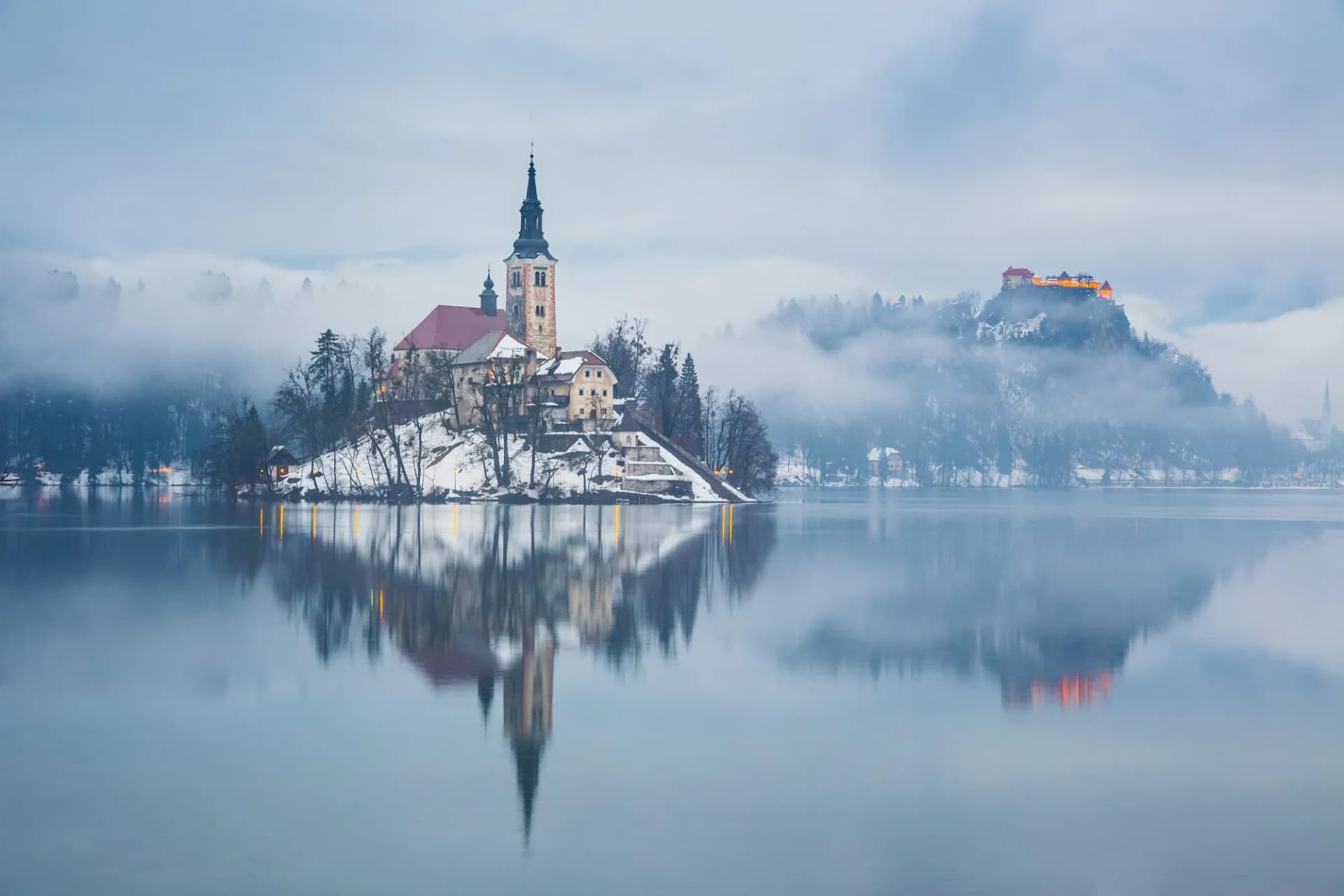 Church on island and castle on hill reflected in Lake Bled with winter fog and snow.