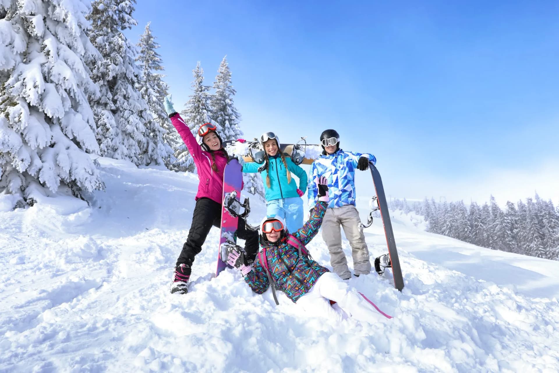 Four snowboarders pose in deep snow on a sunny mountain slope with snow-covered pine trees.