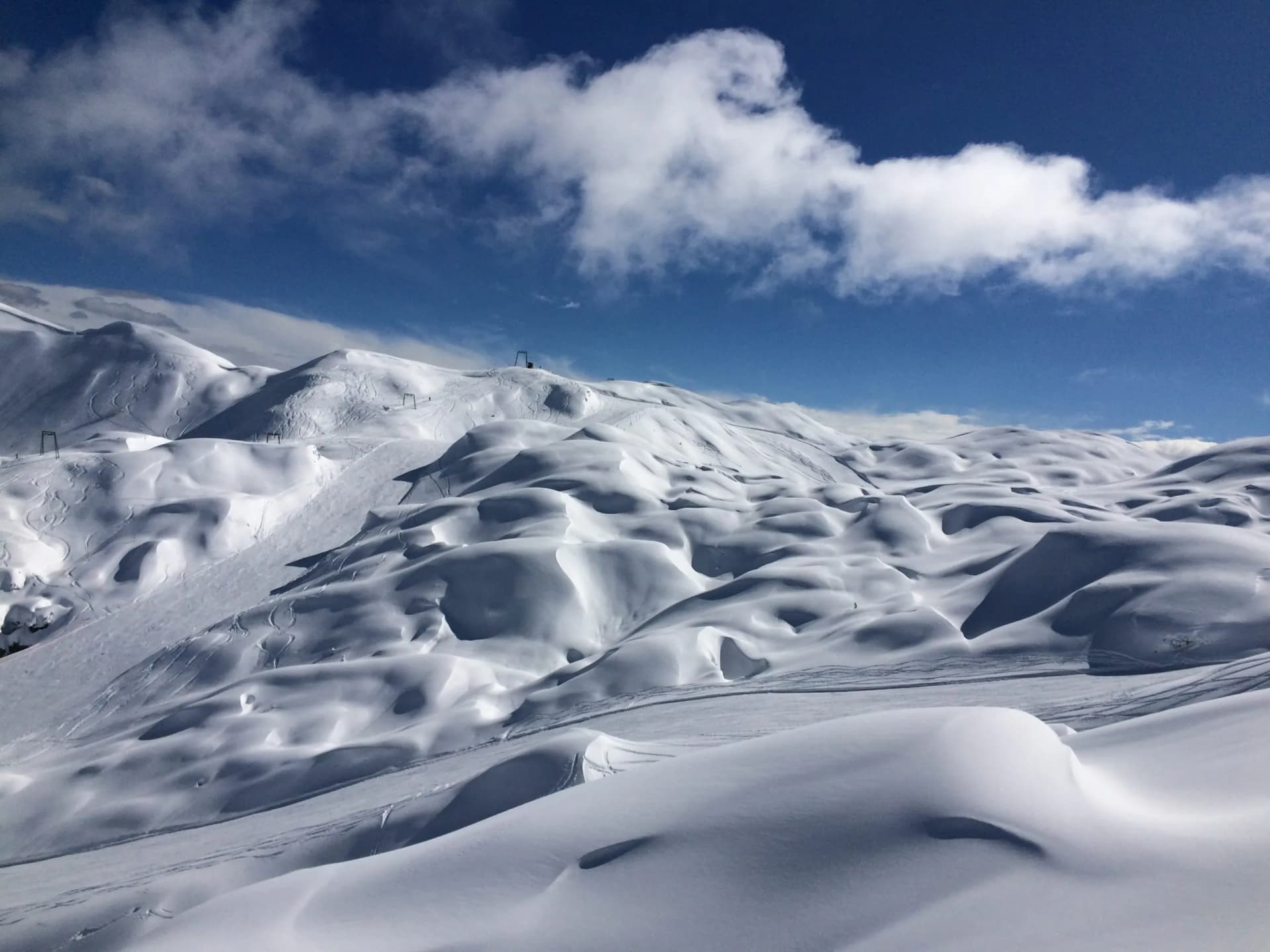 Snow-covered mountain terrain with ski tracks under a bright blue sky with white clouds.