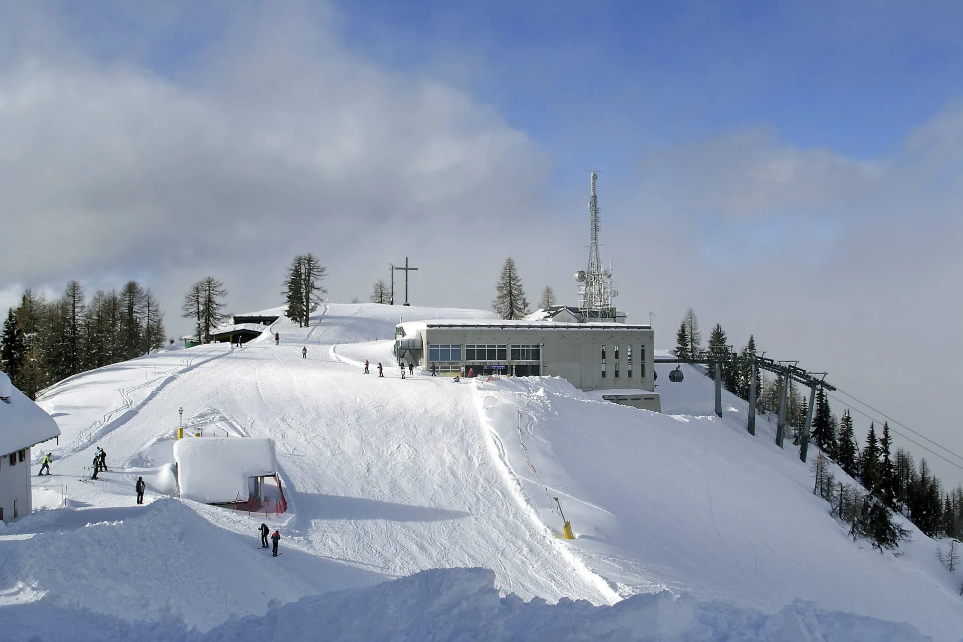 Skiers on snowy slope near modern building, ski lift, and summit cross at Monte Lussari.