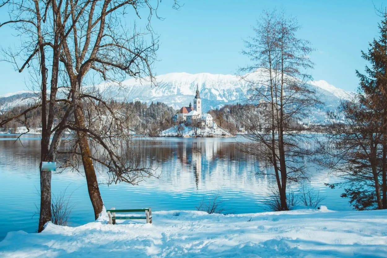 Lake Bled island church in winter with snow-covered mountains reflected in the blue water.