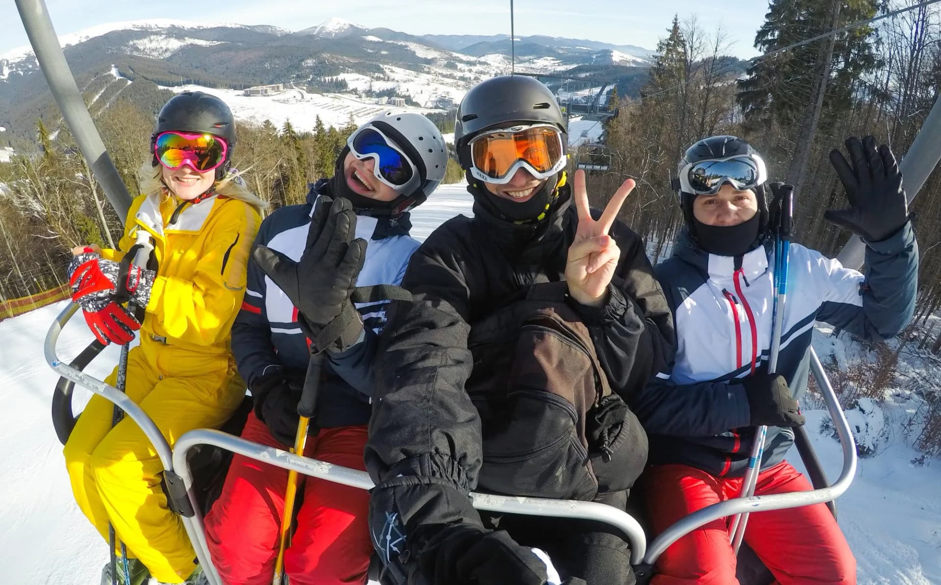 Four skiers on a chairlift waving with snowy mountains in the background