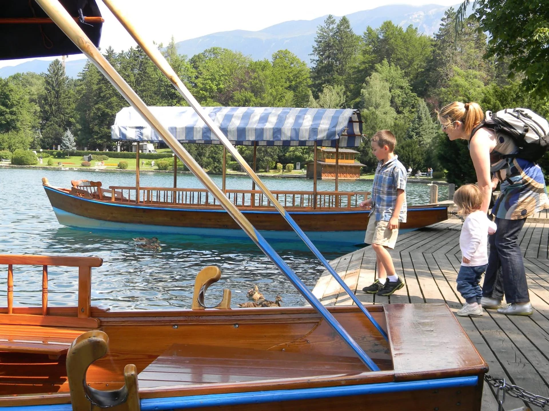 Pletna boat docked on water with family boarding, mountains and forest in background.