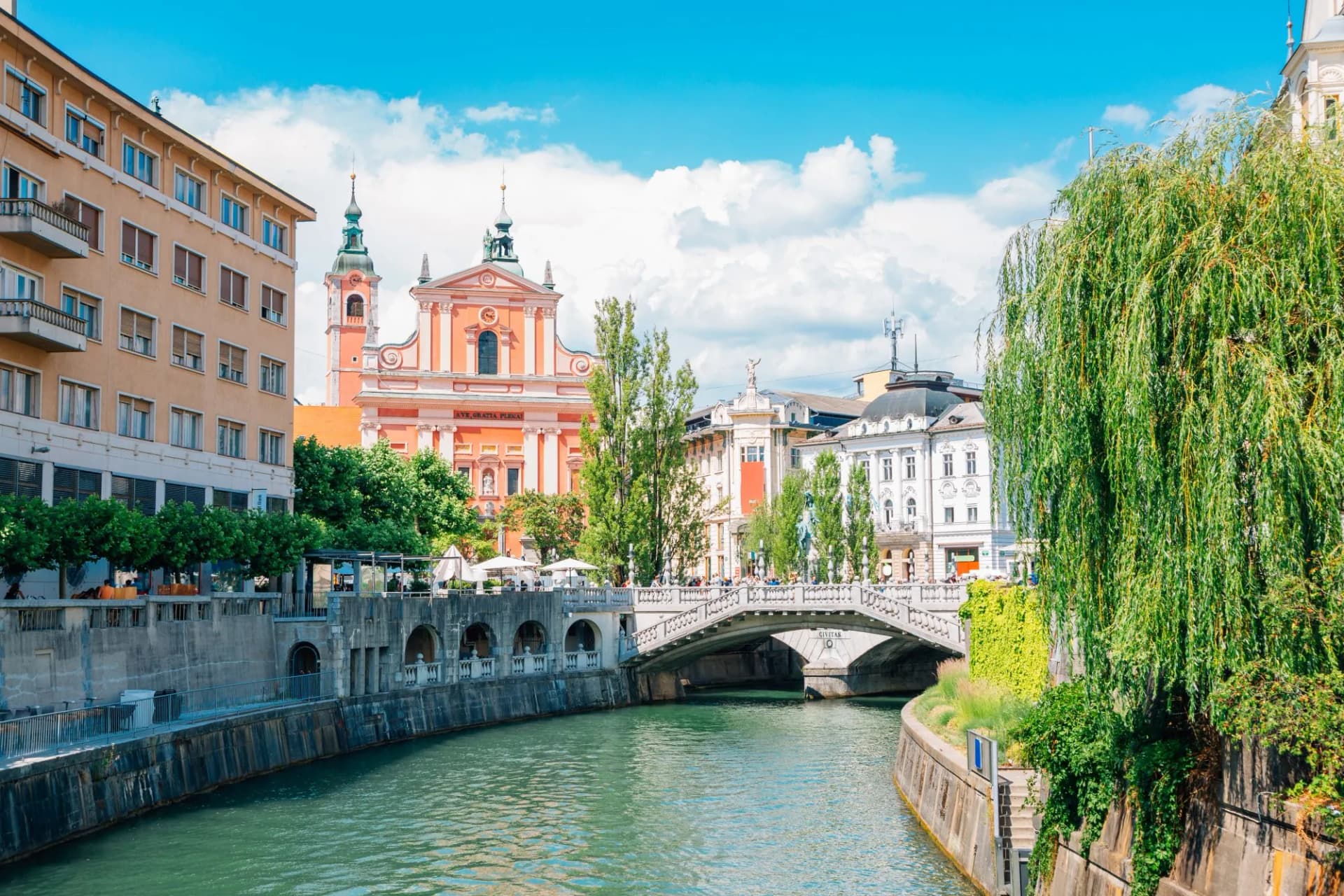 River flowing under a bridge past pink church and buildings in Ljubljana, Slovenia.