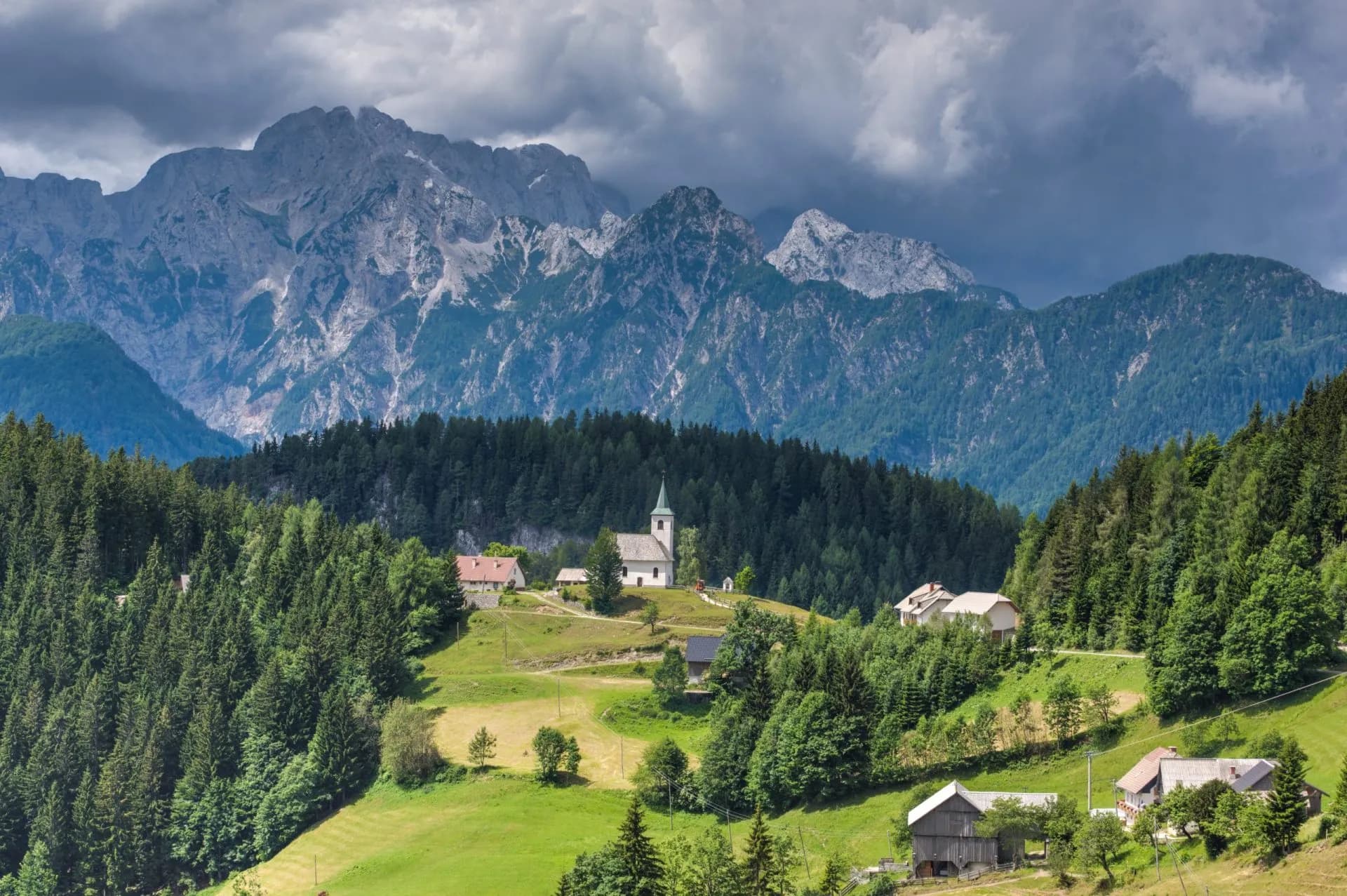 Panoramic view of a small village with a church nestled in green hills below rugged mountains above Logar Valley.