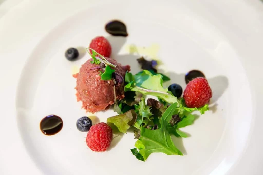 Gourmet meat tartare with raspberries, blueberries, mixed greens, and balsamic reduction on white plate.