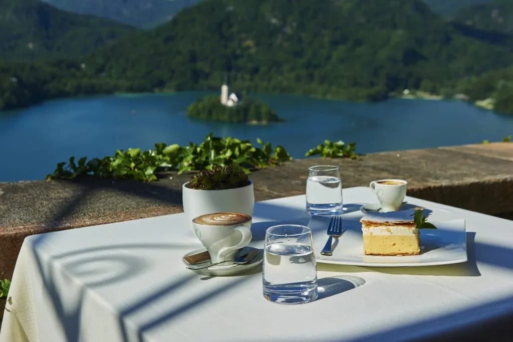 Cafe table with cream cake, coffee, and water overlooking Lake Bled island and mountains.