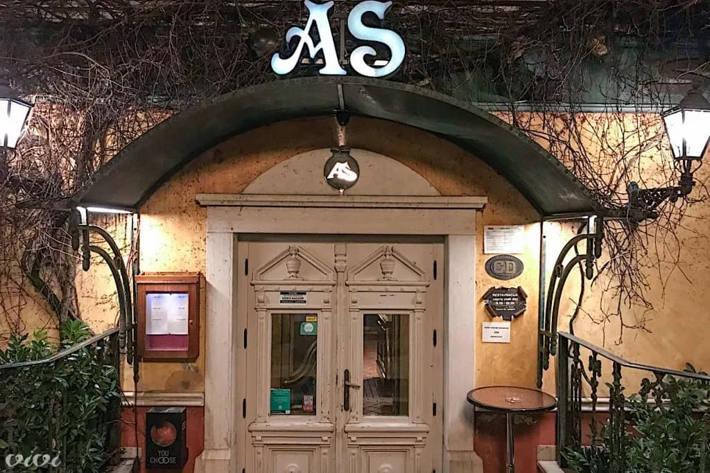 Restaurant entrance with illuminated "AS" sign, double doors, and bare vines overhead at night.