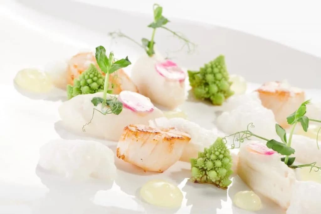 Seared scallops with white puree, Romanesco broccoli, radish, and pea shoots on a white plate.