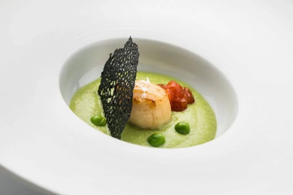 Seared scallop on green pea puree with black crisp and whole peas in a white bowl.