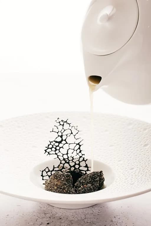 Gourmet dessert with dark crumbles, lace crisp, and liquid poured from a white teapot.