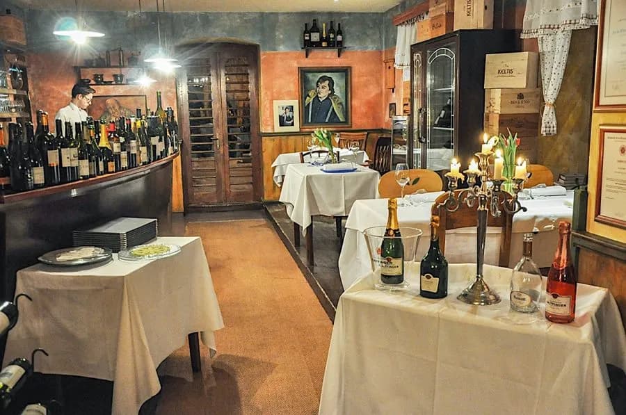 Cozy restaurant interior with wine bottles, white tablecloths, and lit candelabra on tables.