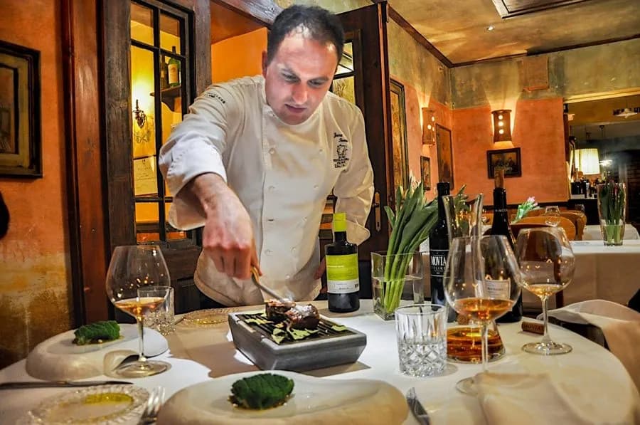 Chef plating food on a small grill at a fine dining table with wine glasses.