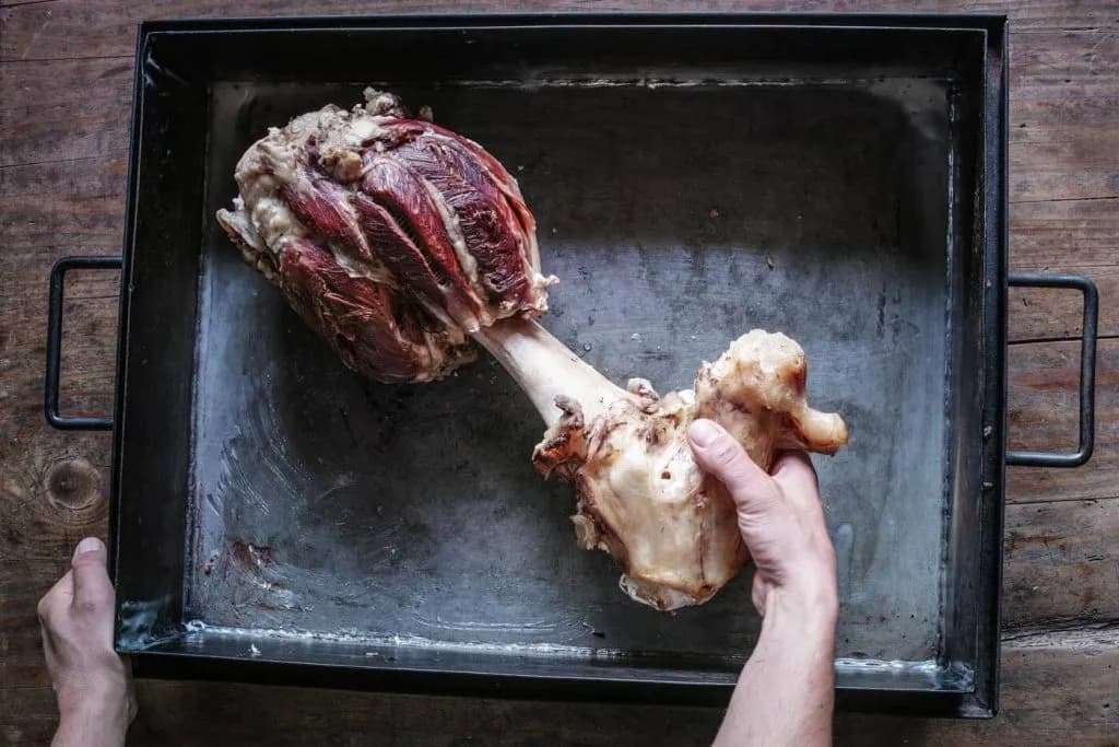 Large piece of raw meat on the bone being placed in a metal roasting pan on wood.