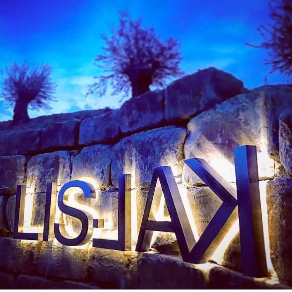 Illuminated "LISJAK" sign mounted on a stone wall against a dark blue twilight sky with trees.