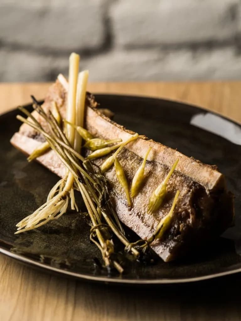 Smoked bone marrow dish with greens and small peppers on a dark plate in a bistro setting.