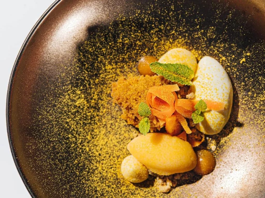 Gourmet dessert with yellow powder, quenelles, mint, and candied fruit on a dark plate.
