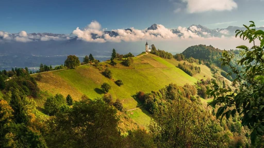Church on the top of Jamnik hill with green slopes and distant mountains under clouds