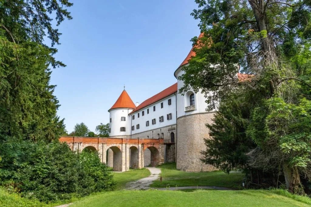Brežice Castle with white walls, red roofs, and arched stone bridge surrounded by lush green trees.