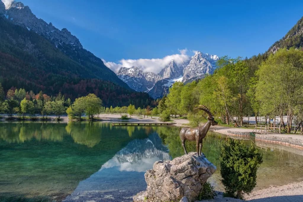 Bronze ibex statue by clear alpine lake with snow-capped mountains and green trees