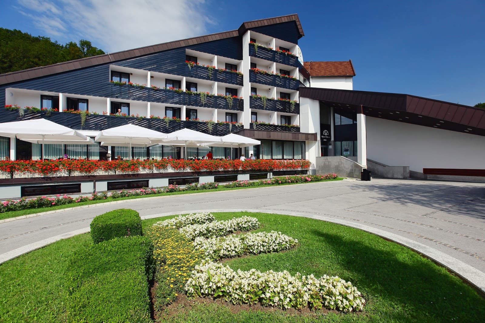 Hotel Breza exterior with balconies, red flowers, white umbrellas, and manicured lawn.