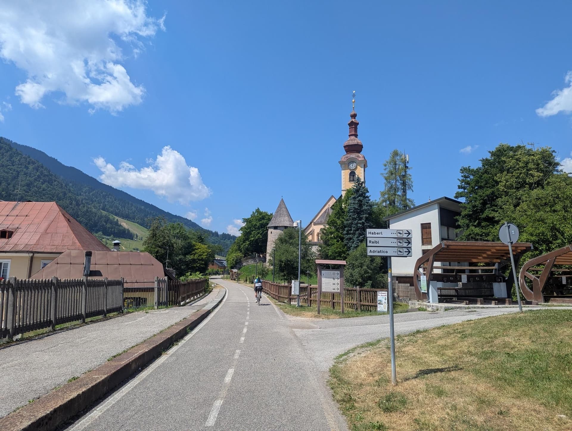 Cyclist on a paved trail leading toward a historic church tower in an alpine village.