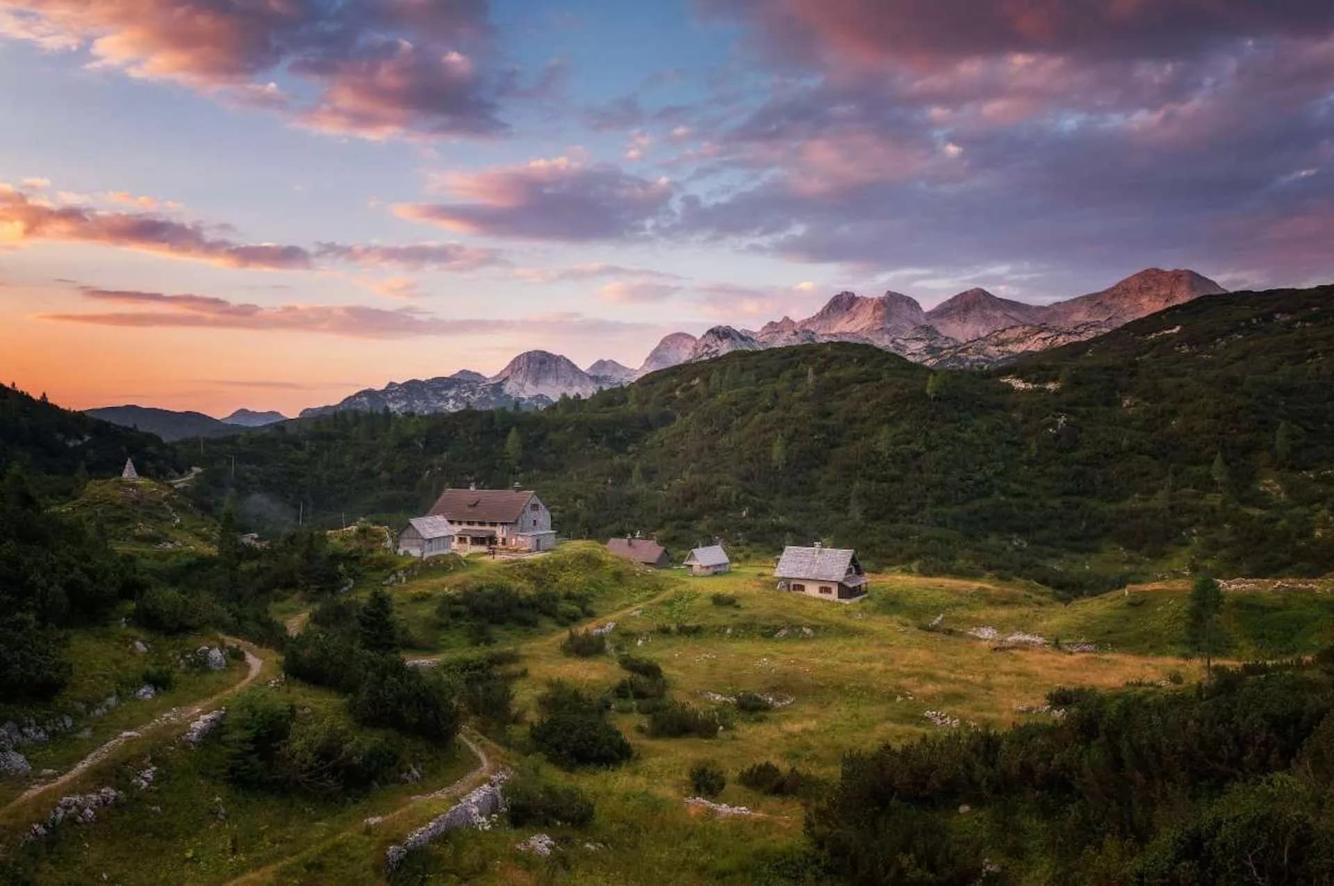 Mountain huts in a green alpine meadow below rugged peaks at sunset, Komna Plateau.