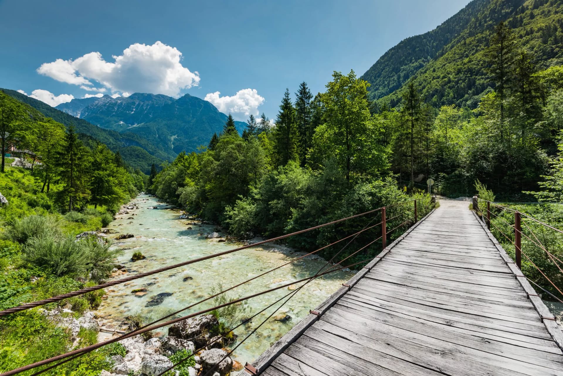 Wooden suspension bridge over the turquoise Soca River in Slovenia with forested mountains.