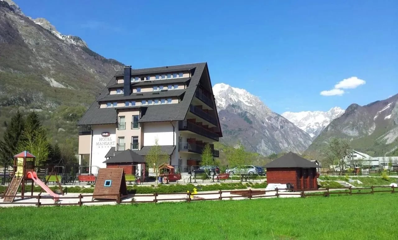 Hotel Mangart with playground in front of snow-capped mountains under a blue sky.