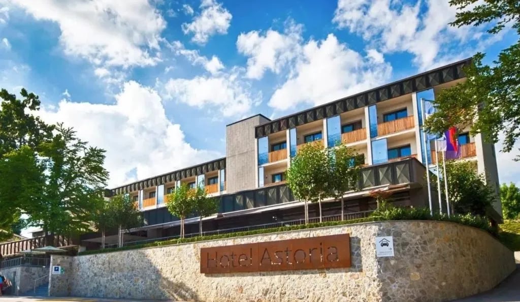 Hotel Astoria building facade with stone retaining wall and green trees under a blue sky.