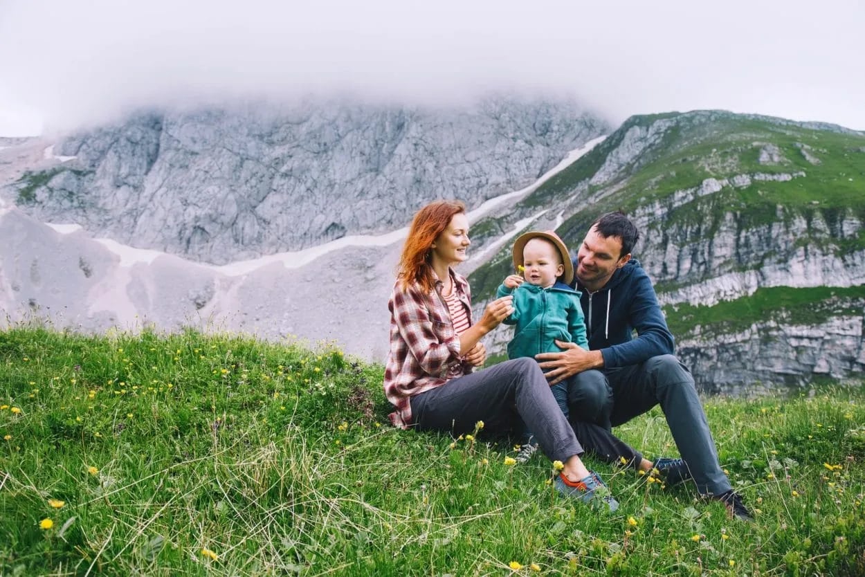Family relaxing on grassy slope with wildflowers below misty, rocky alpine mountains.
