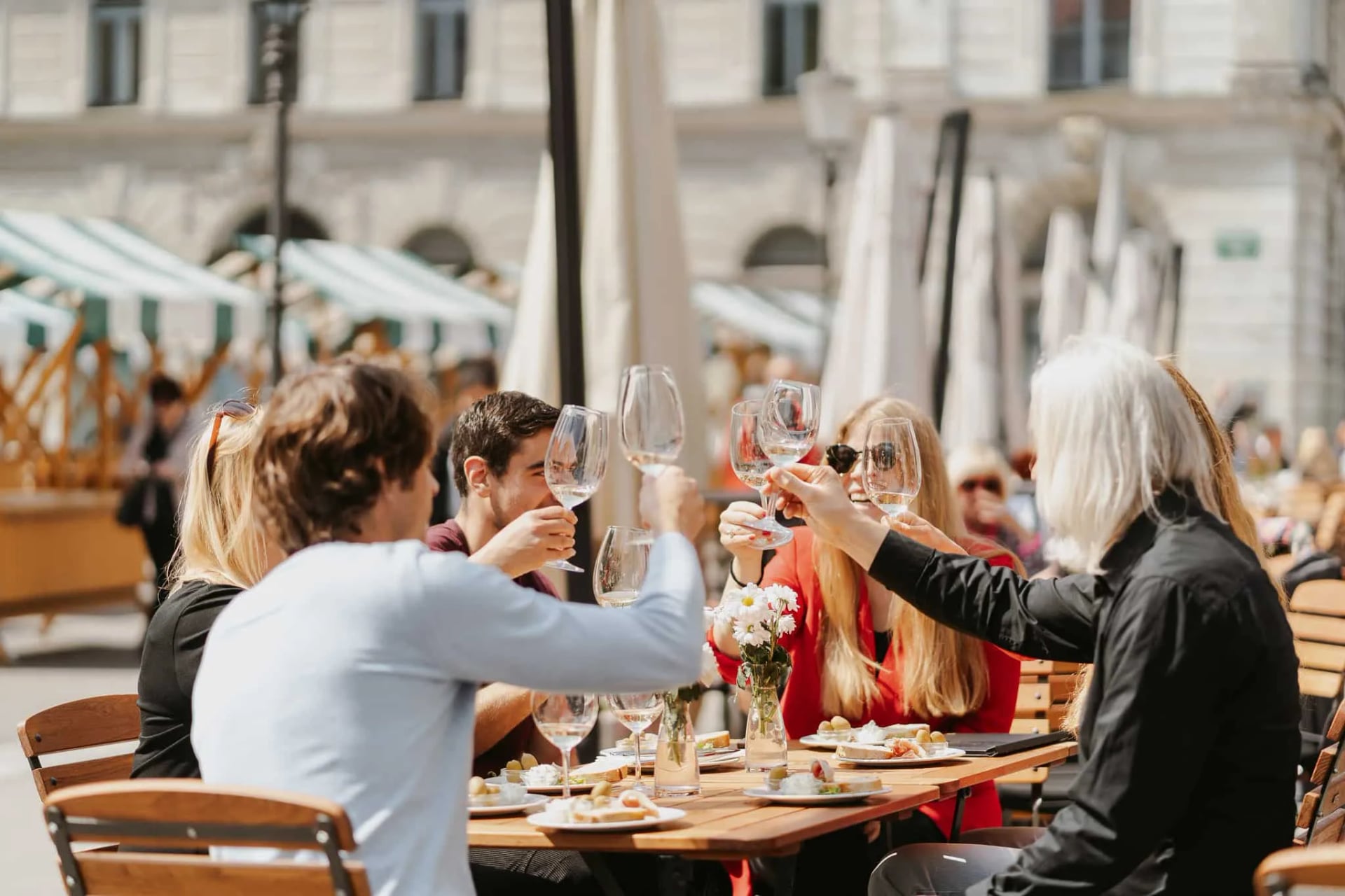 Group toasting with wine glasses at outdoor cafe table with food and flowers.
