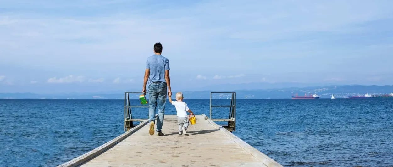 Father and child walking on pier toward blue sea with distant cargo ships and mountains.