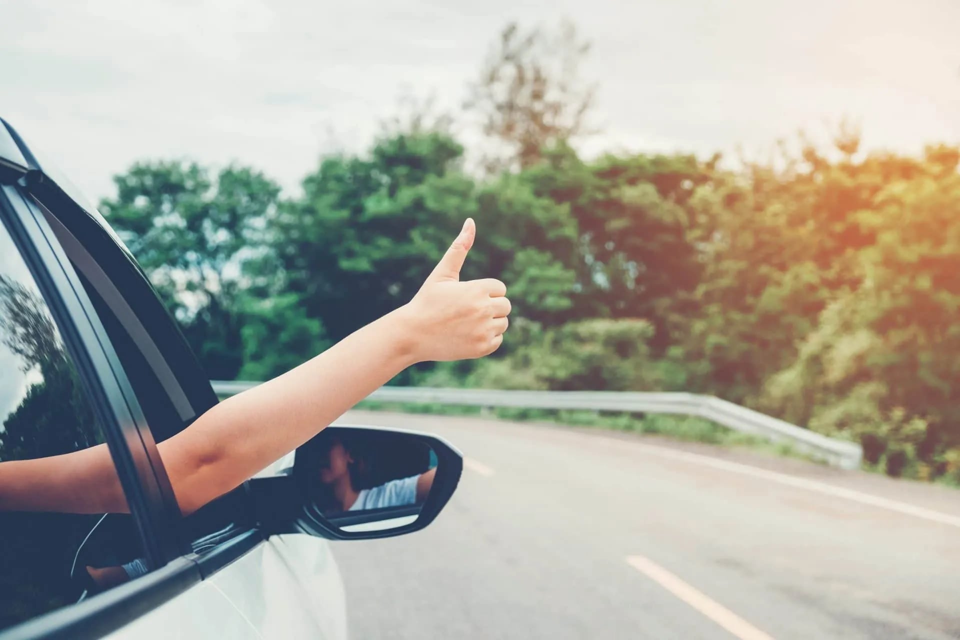 Arm giving thumbs up out of car window while driving on road with trees