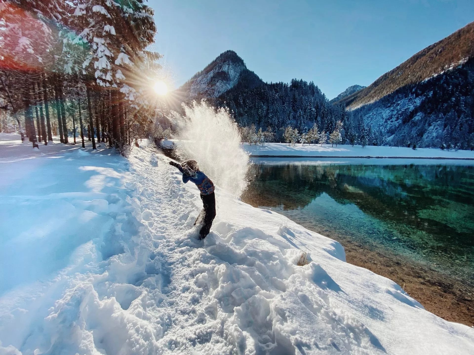 Child throwing snow by turquoise alpine lake with snow-covered mountains in winter, Slovenia.