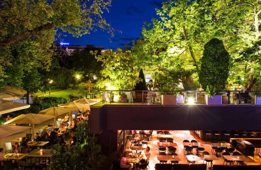 Outdoor aperitivo restaurant dining area with multiple levels illuminated under lush green trees at night.
