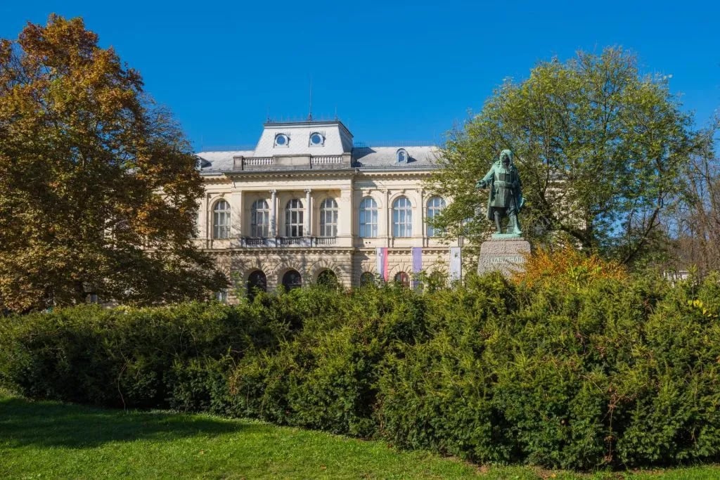 Ljubljana National Museum building with statue in foreground garden under clear blue sky.
