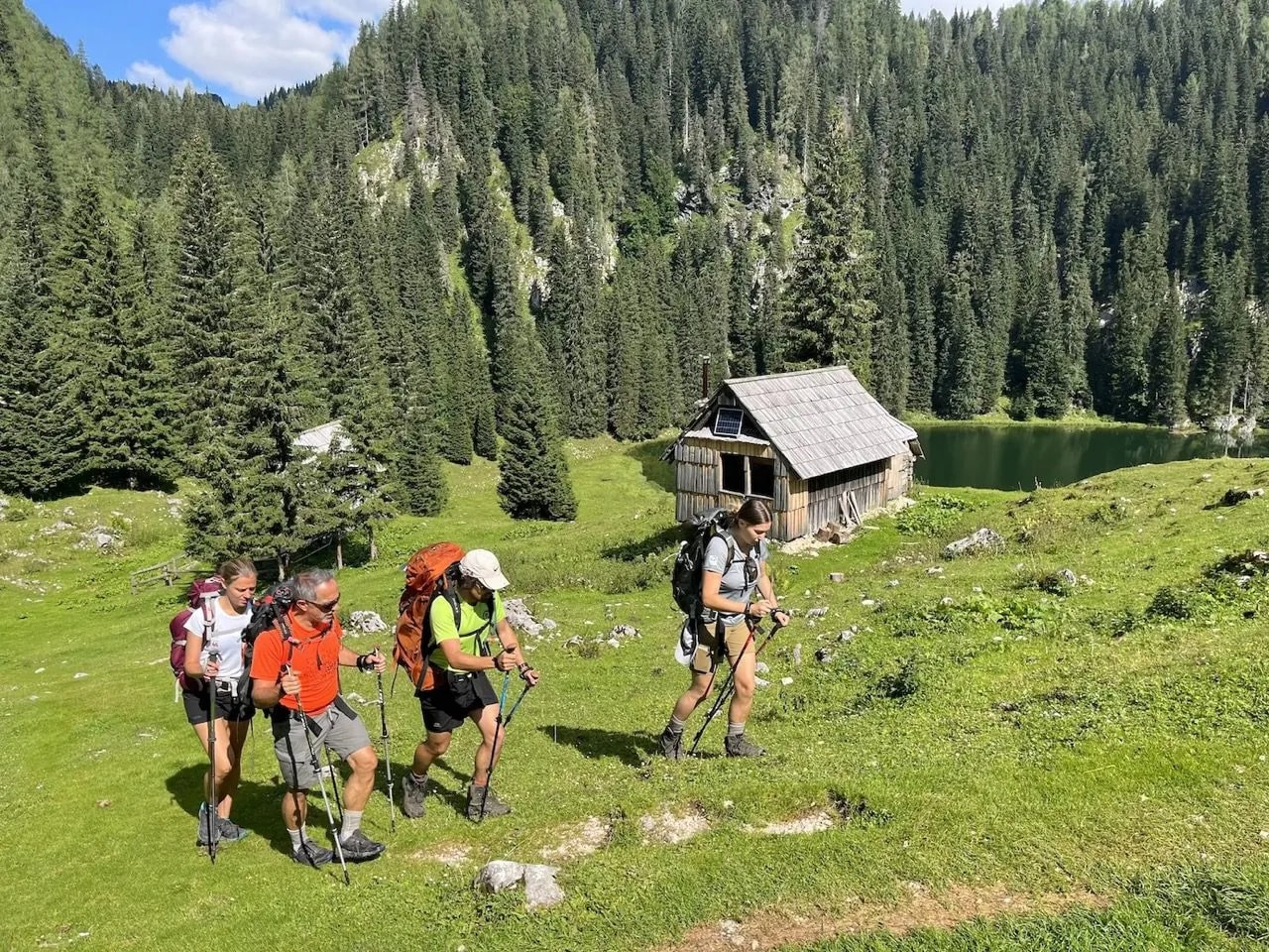 Hikers with backpacks and poles walk past a wooden cabin near a lake in a forested mountain area.
