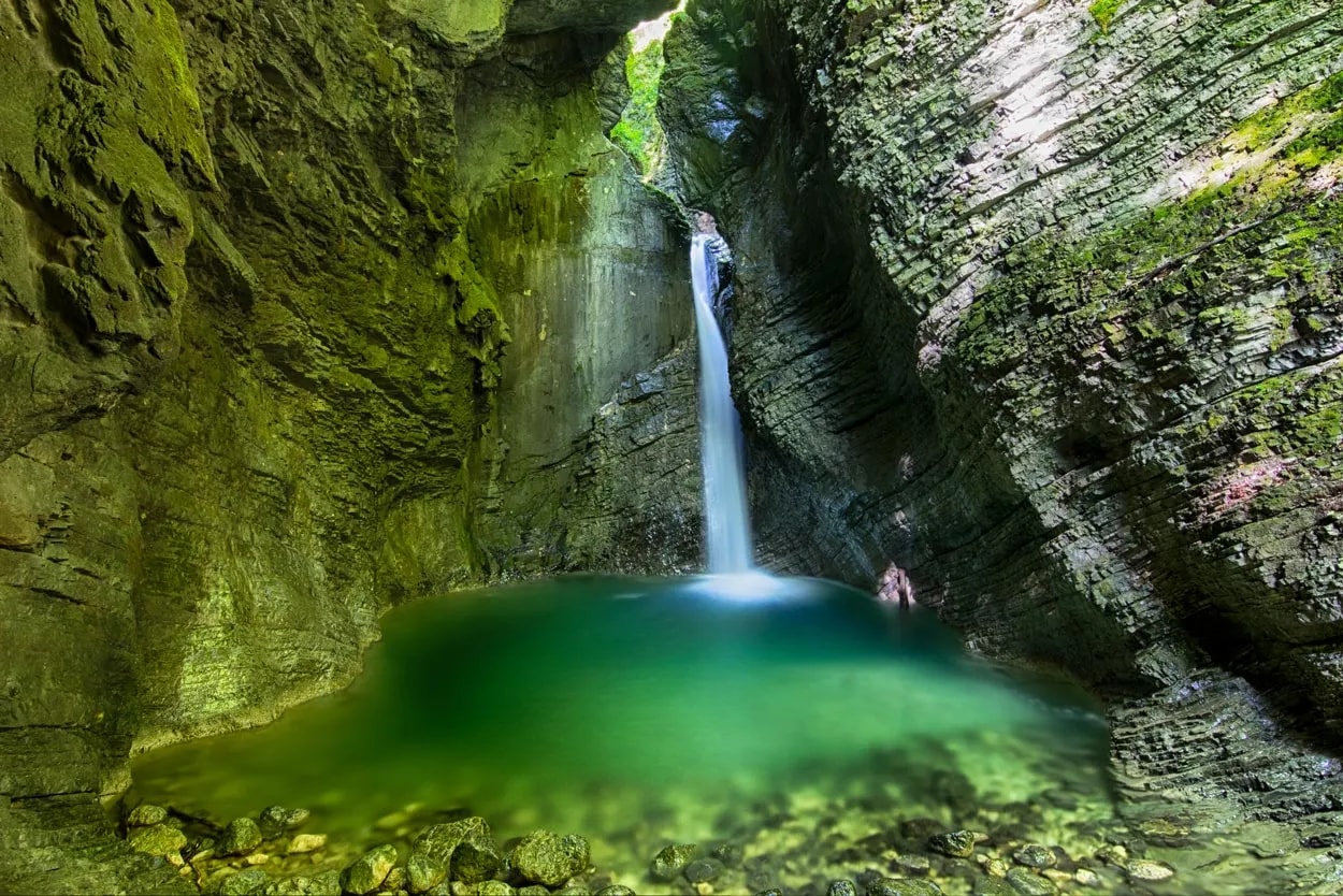 Waterfall cascading into a green pool inside a mossy, narrow rock canyon at Kozjak Waterfall.