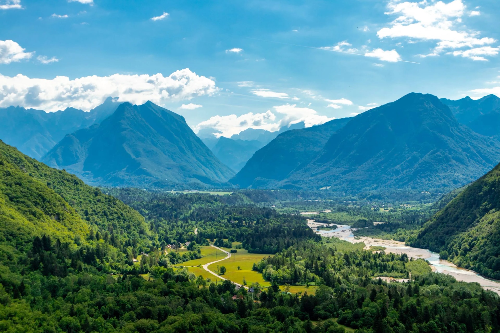 Lush green valley with a winding river, surrounded by steep, blue mountains under a cloudy sky.