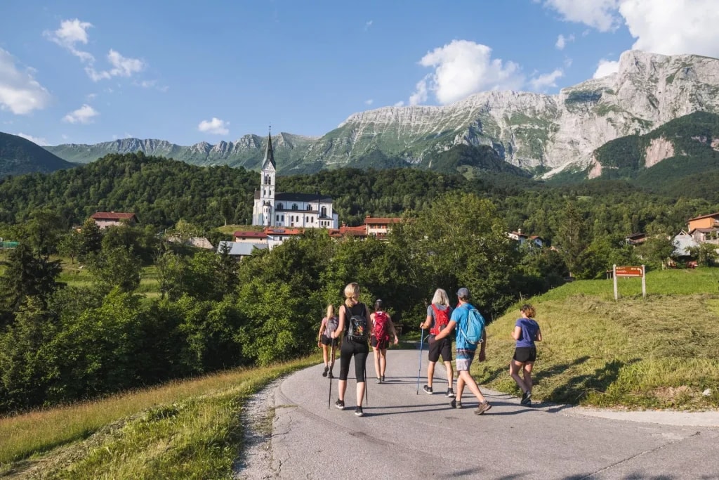 Hikers walking toward a village with a white church under Mt. Krn in Drežnica.