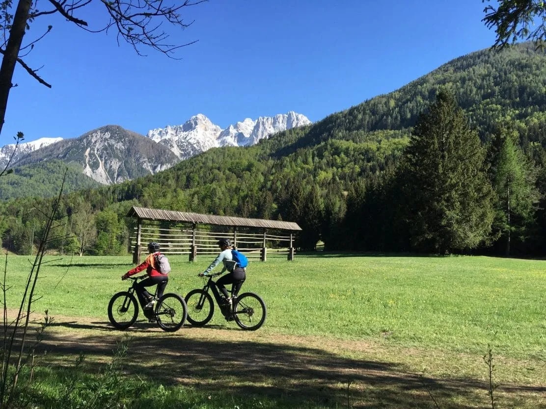 Two cyclists riding mountain bikes across a grassy field with snow-capped mountains in the background.