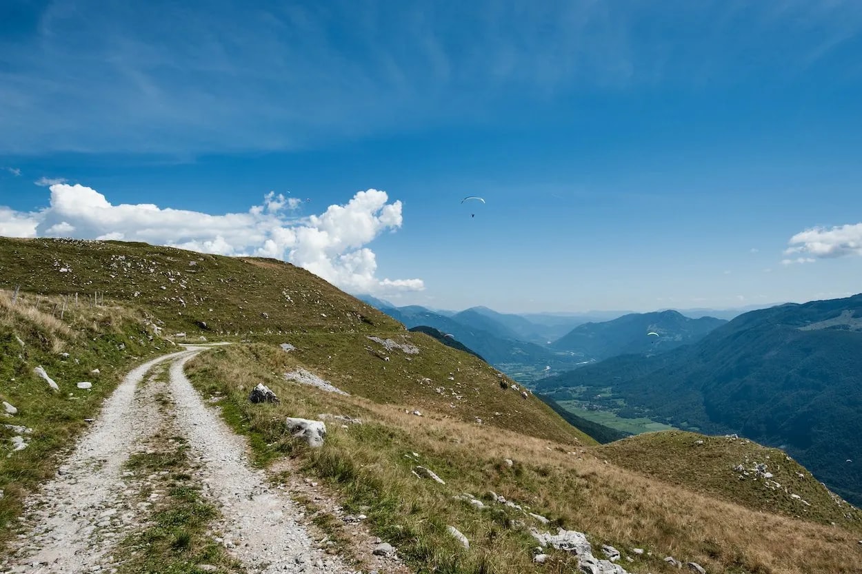Dirt road on grassy mountain slope with paragliders over a deep valley.