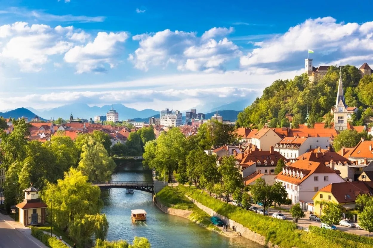 Ljubljana city view with river, castle on hill, and distant mountains under blue sky.